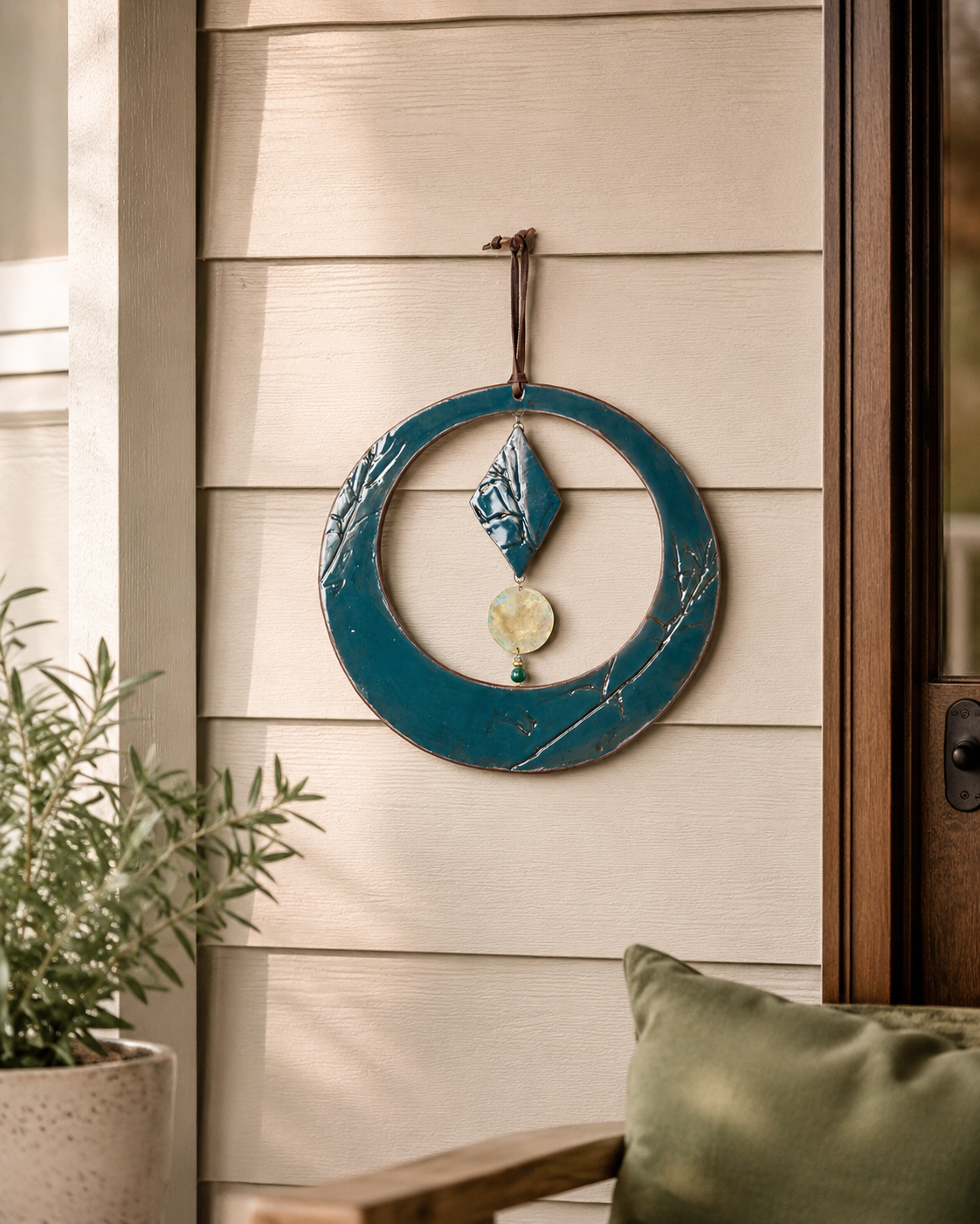 A teal blue round ceramic wall hanging with a hanging loop, displayed on a beige exterior house siding door next to a potted plant. FTF 0726