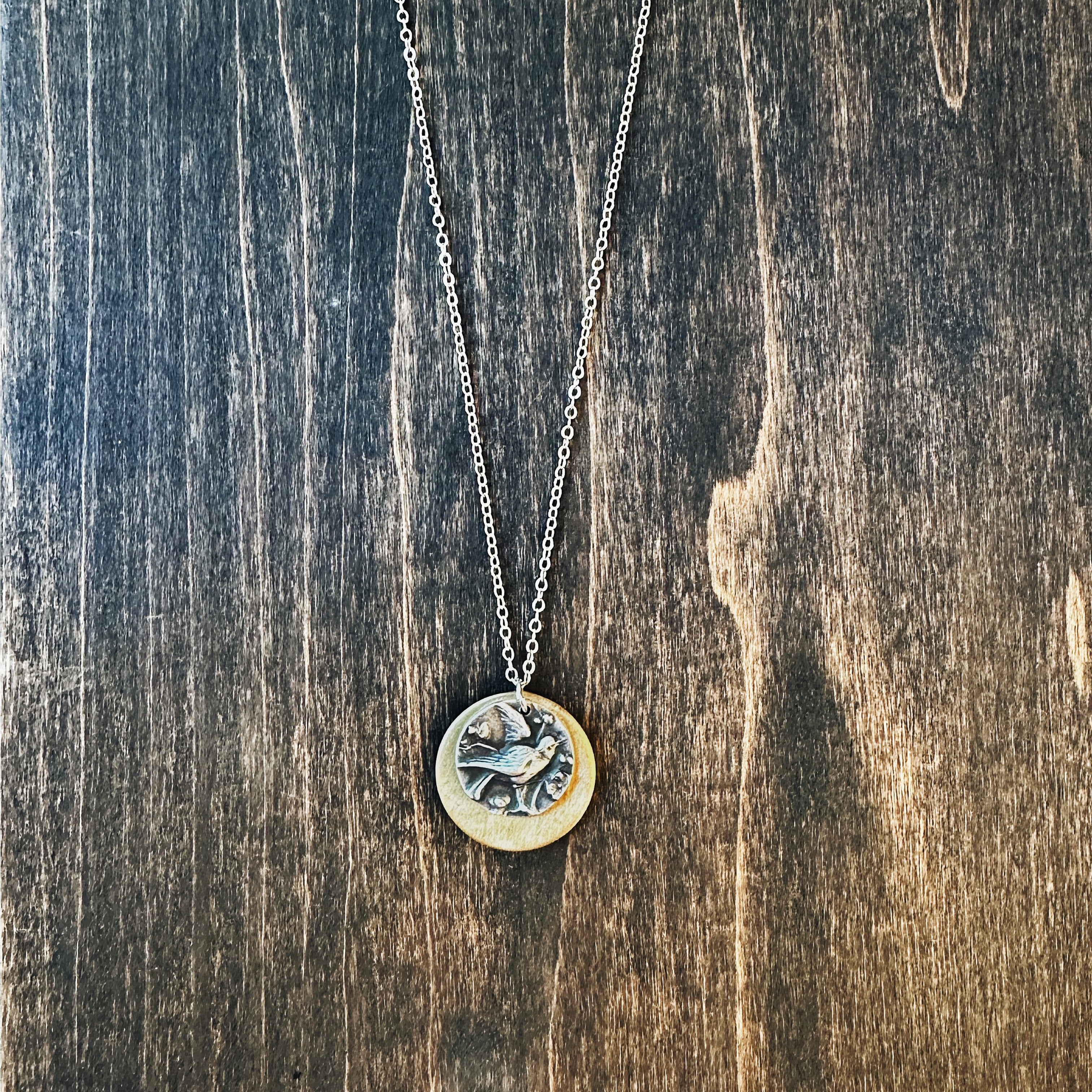 Birds of Flutter mixed metal charm necklace on dark wood surface, silver bird charm and bronze disc, by Jester Swink