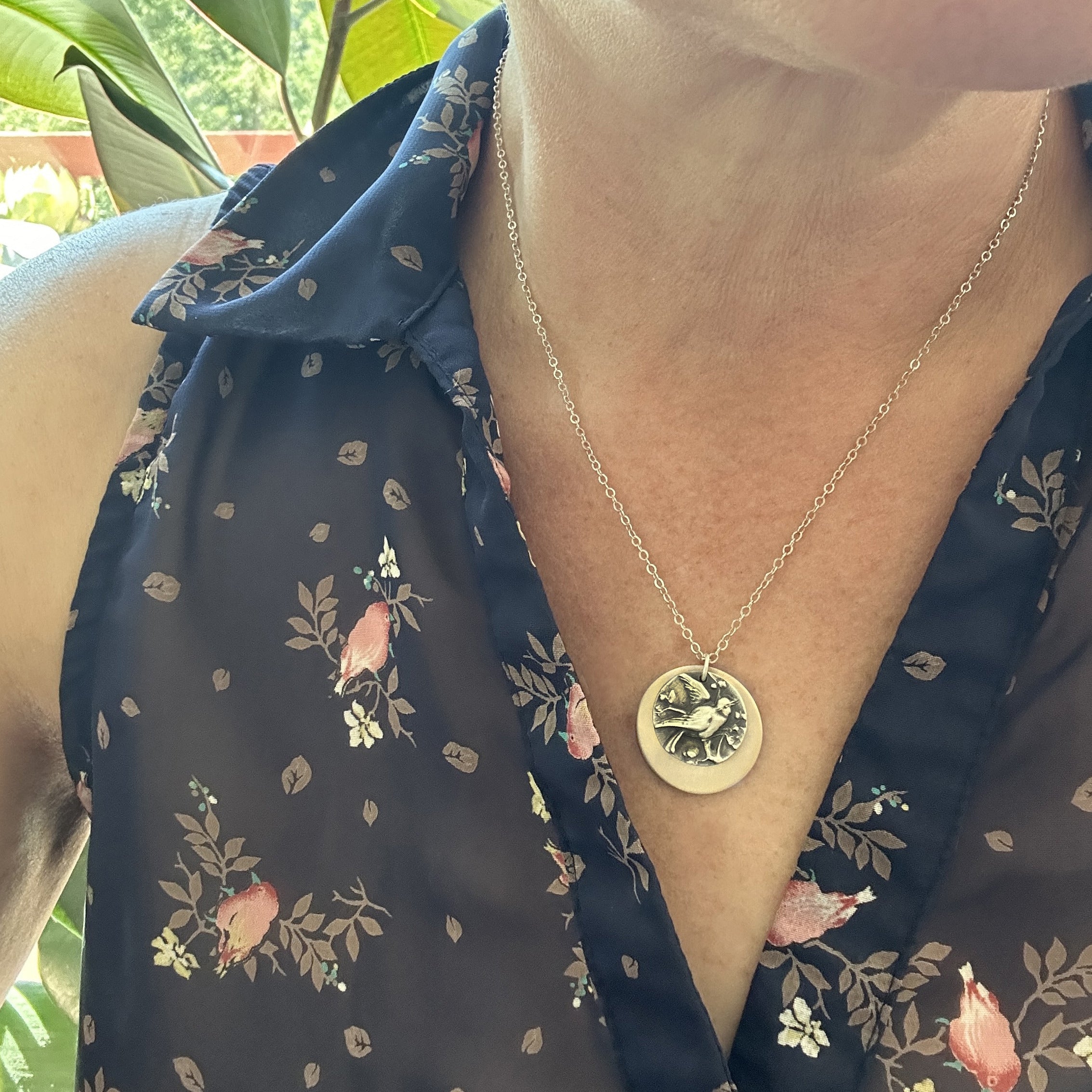Birds of Flutter mixed metal charm necklace worn, showing silver bird charm and bronze disc against floral blouse, by Jester Swink