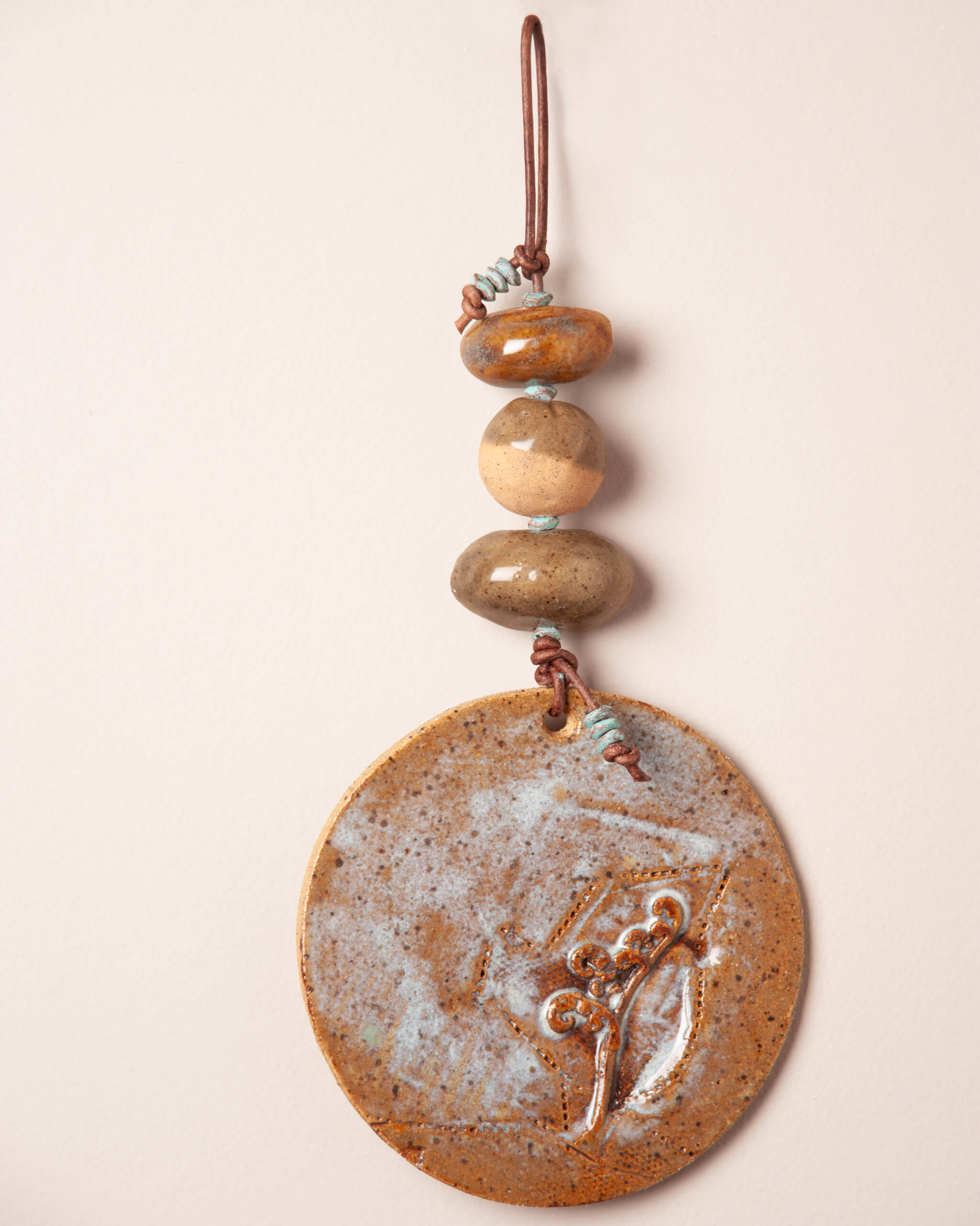 Close up of raised fiddlehead fern impression on sandy ice blue glazed ceramic disc with patinated brass and teal accent beads