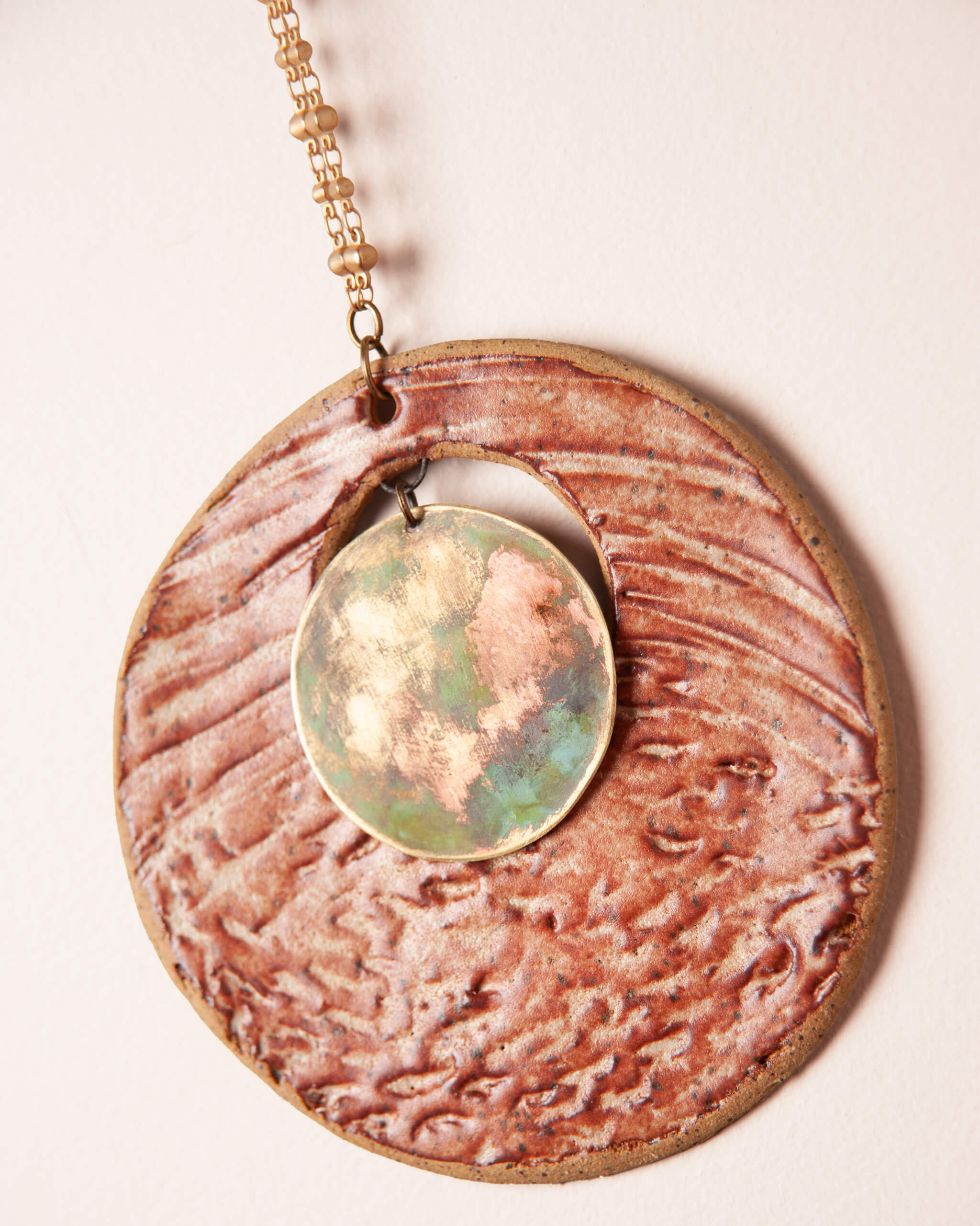 Close up of hand patinated brass disc showing copper gold and green oxidation detail suspended on brass jump ring inside Patina Shore Moon ceramic wall hanging