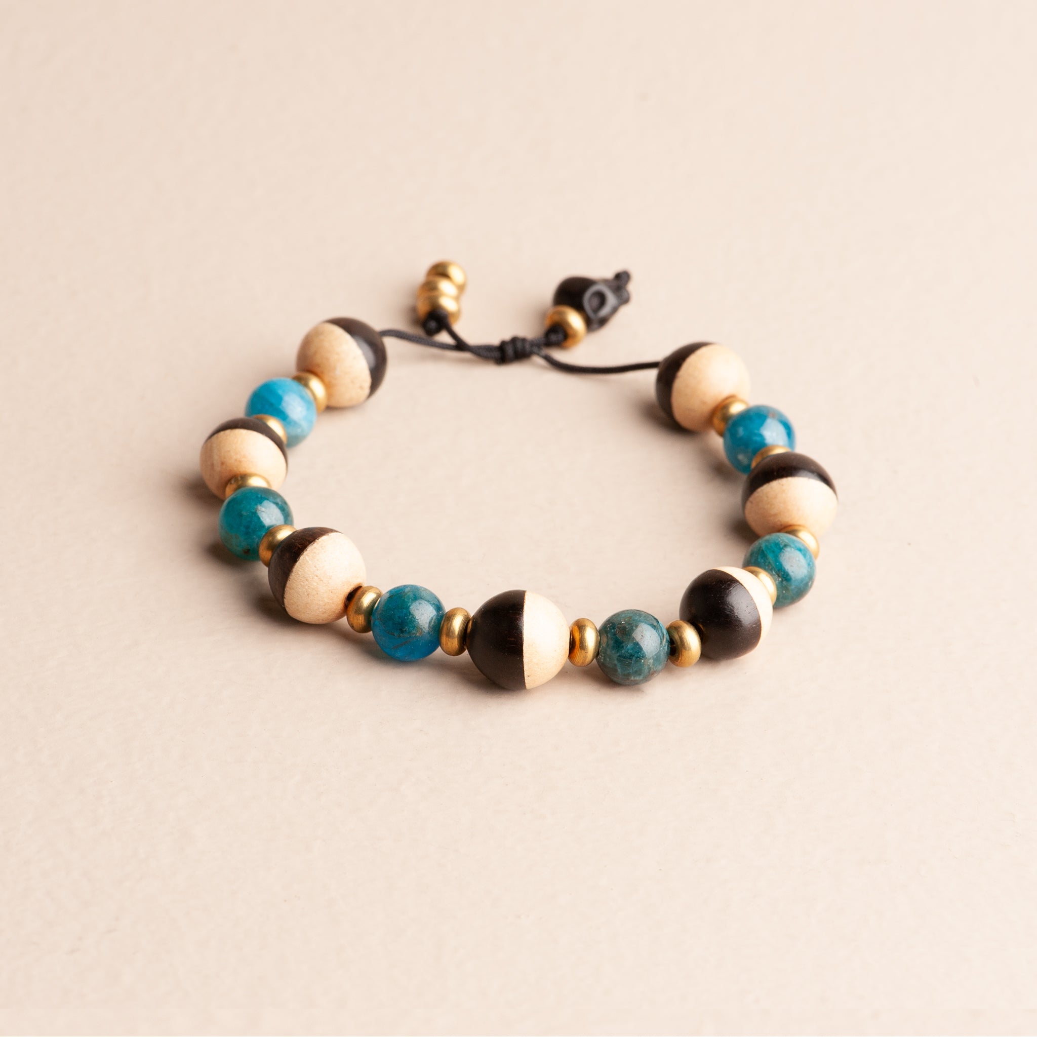 Adjustable nylon cord bracelet featuring bright blue Apatite beads, half-painted black and white wood beads, and brass accents. The dangle detail includes a small black wood skull.