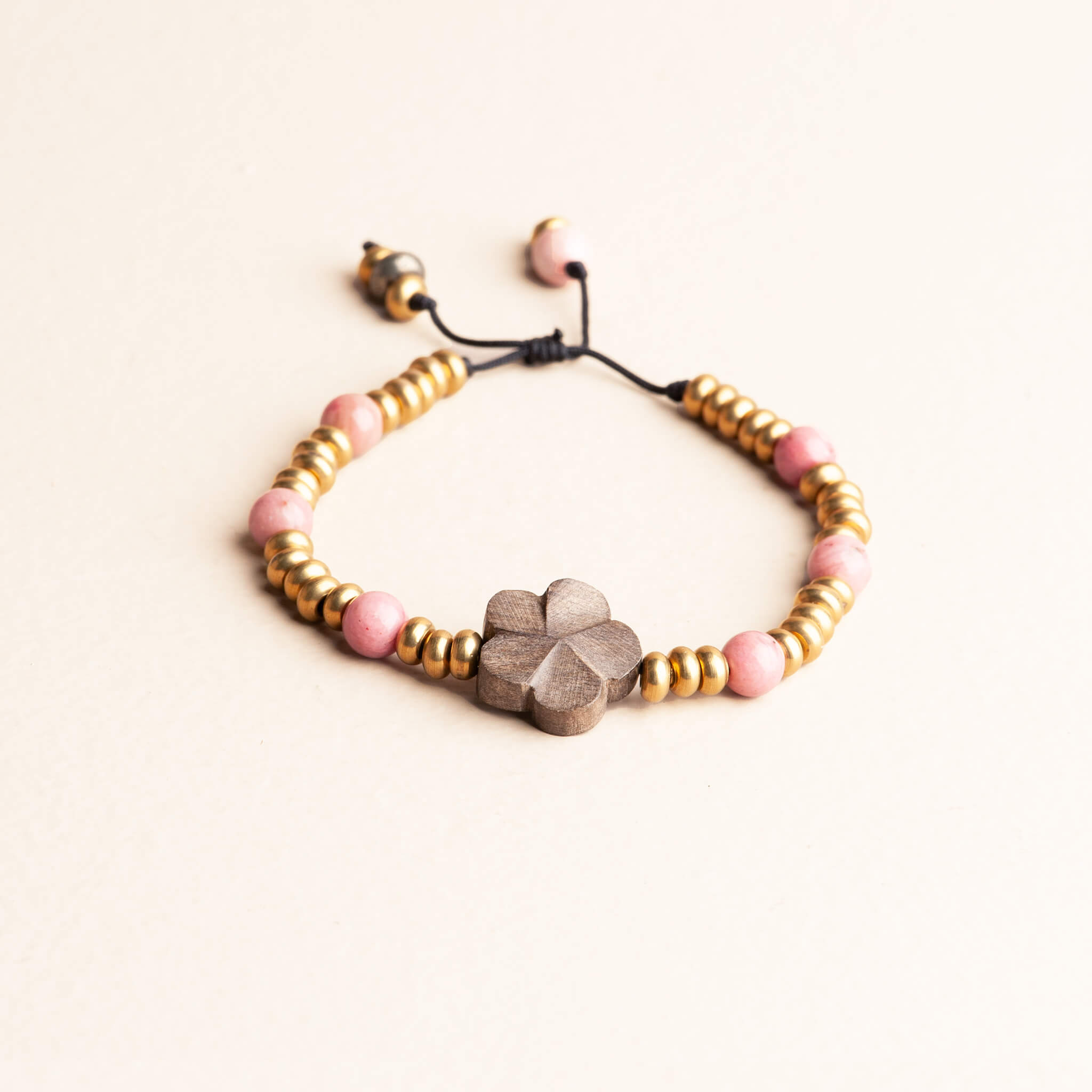Adjustable knotted bracelet featuring pink Rhodochrosite beads, brass spacer beads, and a central grey wood flower focal bead on a dark nylon cord.