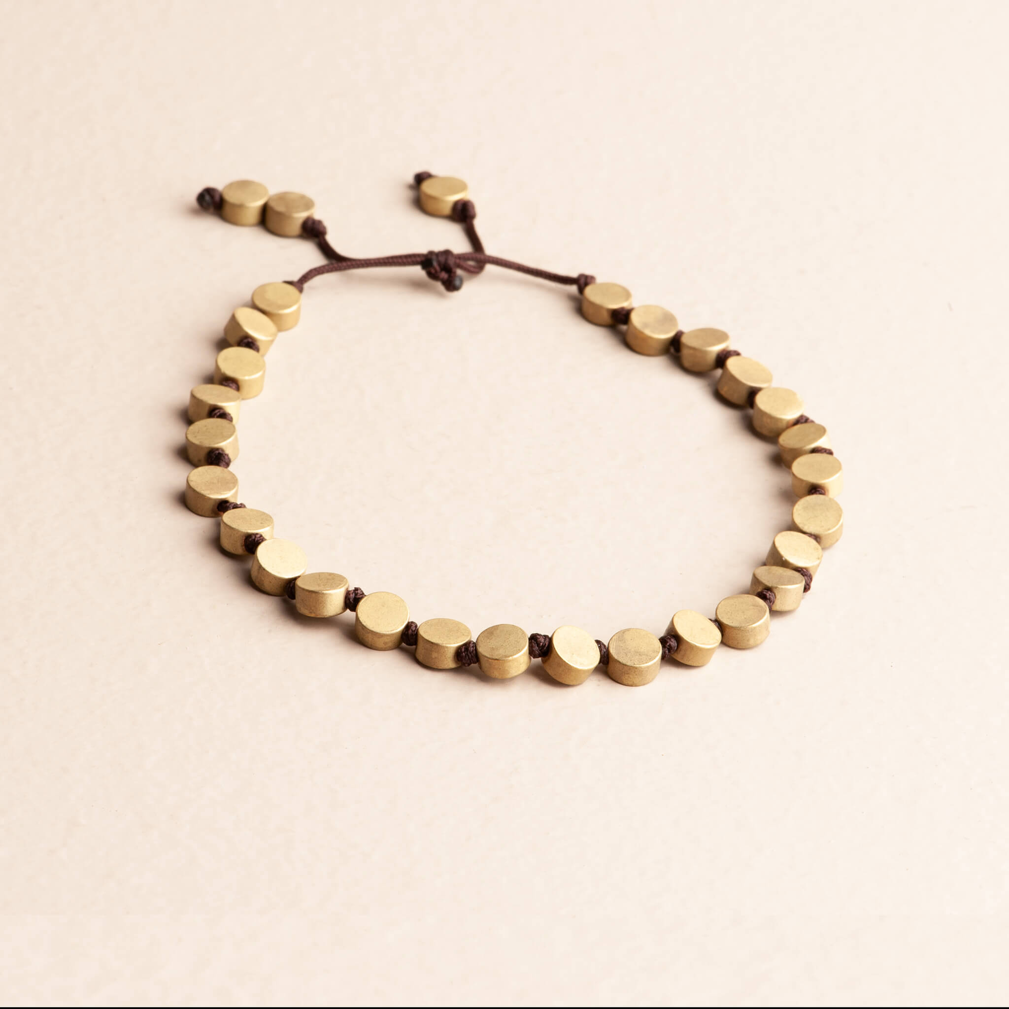 Modern Boho adjustable slider bracelet featuring flat, round solid brass coin beads knotted on a dark brown nylon cord, photographed on a cream background.