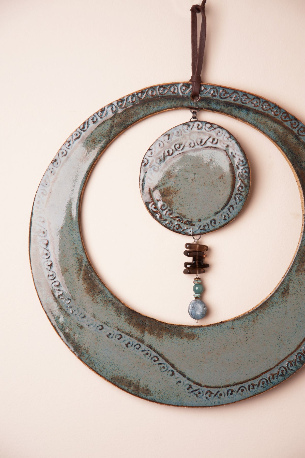 Detail of the smoky topaz, kyanite and aventurine stone dangle below the pressed scroll disc on a large handmade round ceramic wall hanging, kiln fired by Jester Swink, Santa Cruz Mountains California