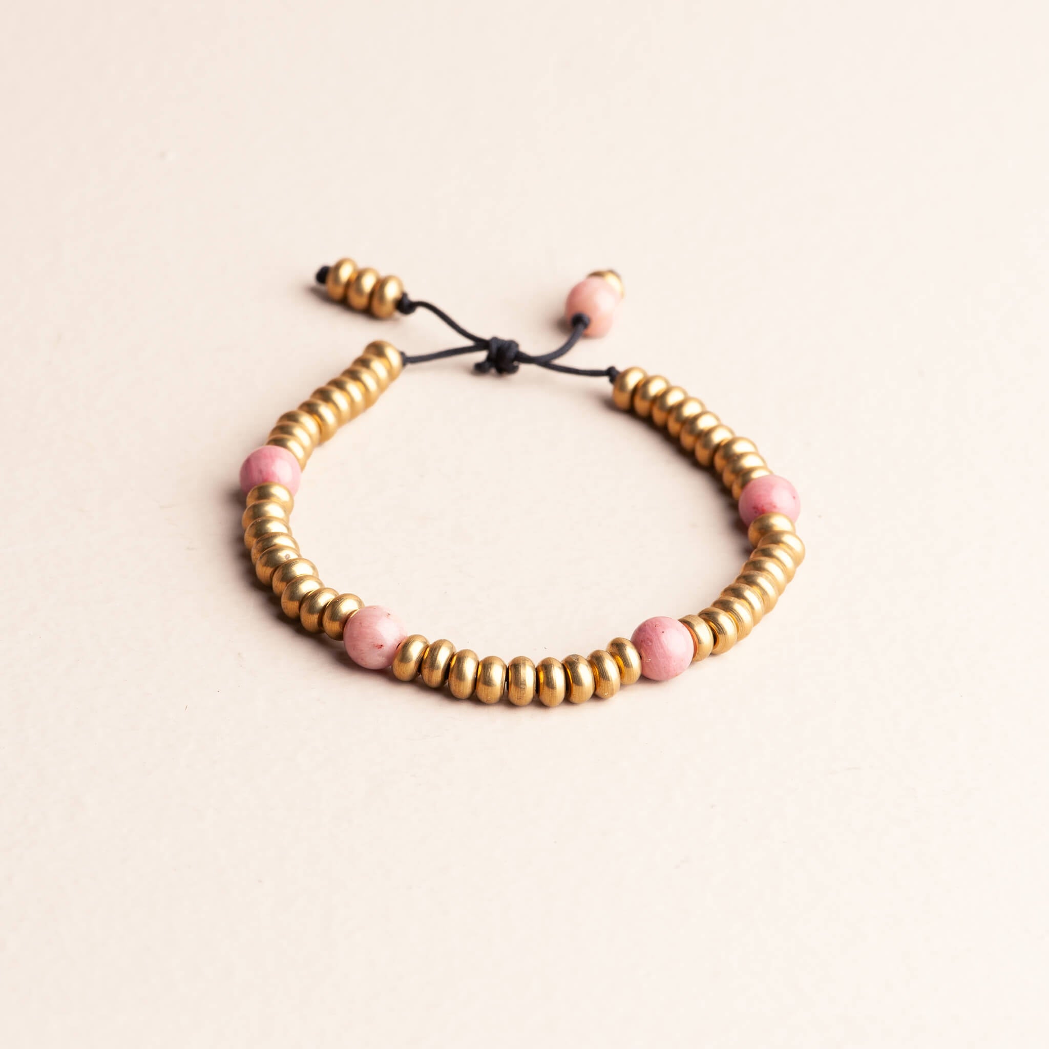 Adjustable knotted bracelet featuring round brass spacer beads and four pink Rhodochrosite beads on a dark nylon cord, photographed on a cream surface.
