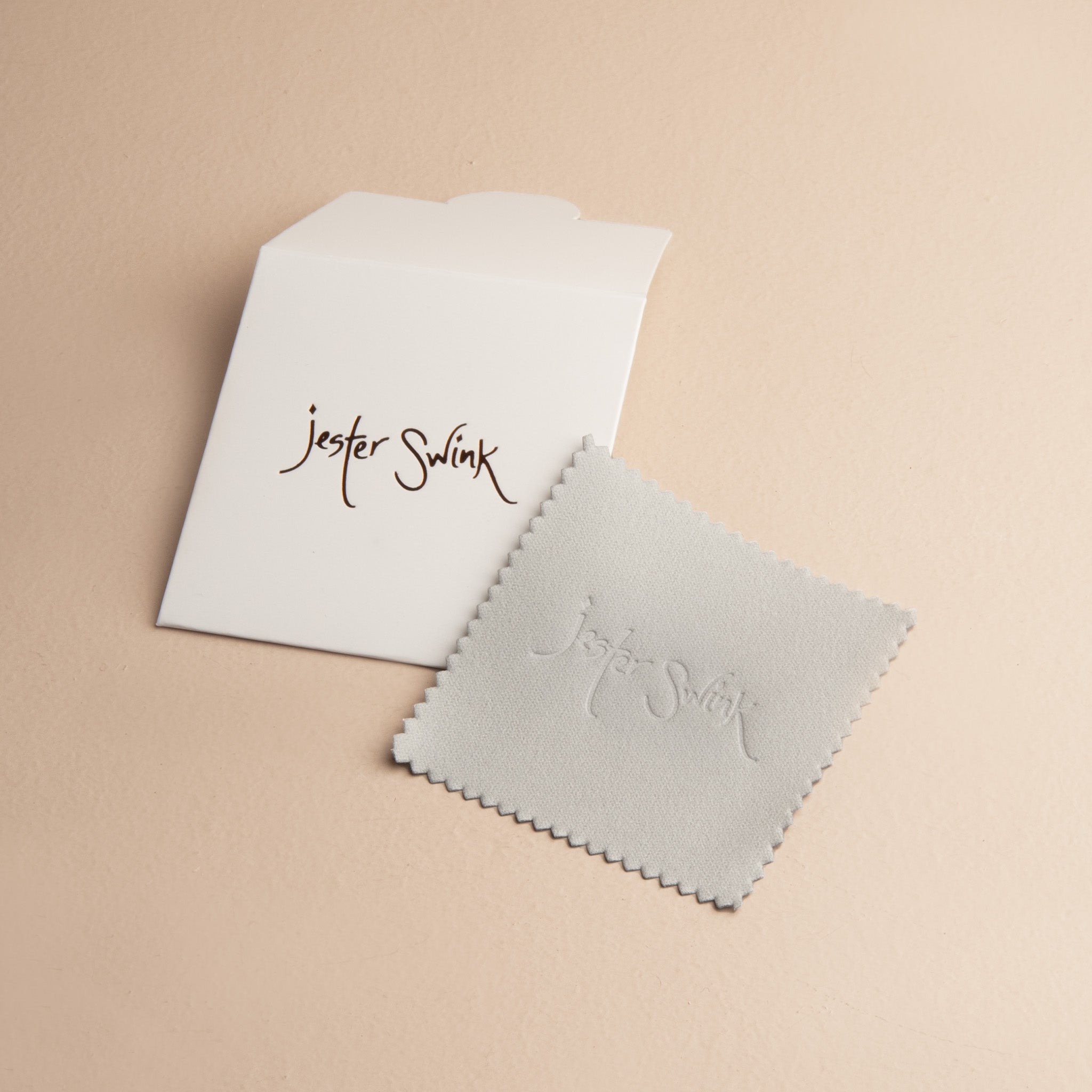 Jester Swink branded travel jewelry polishing cloth laid next to its white protective envelope, showing the scalloped edges.