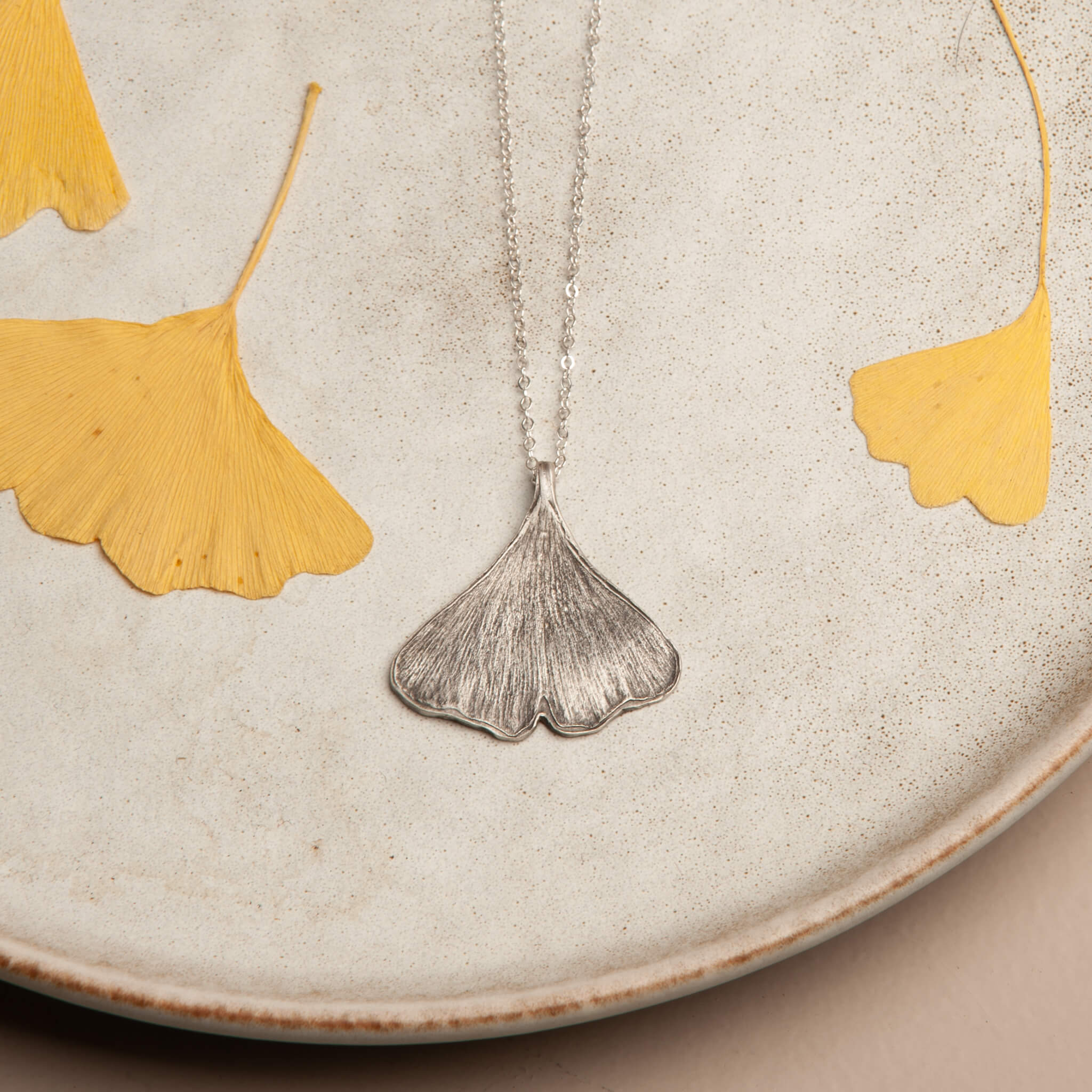 Sterling silver ginkgo leaf pendant inspired by a real ginkgo leaf, shown with natural leaves for scale and texture